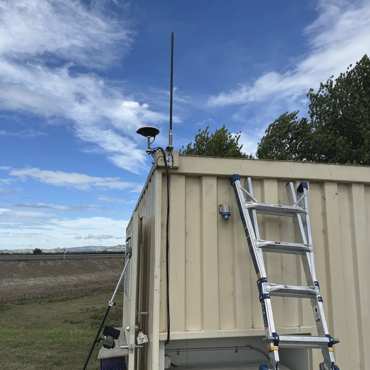 RTK base antenna and UHF radio mounted on the corner of a site container beside a highway, with a ladder up during install