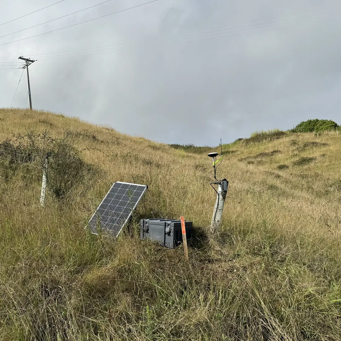 Solar-powered RTK base with GNSS antenna on a fence post at a remote rural hillside site